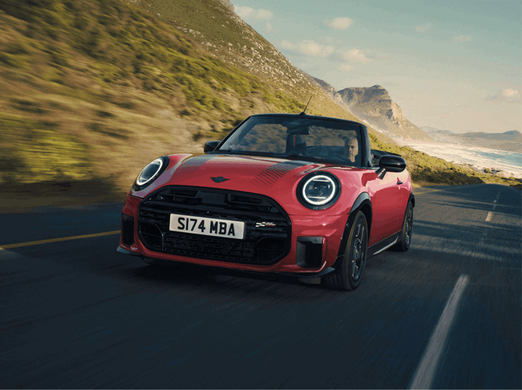 Ein roter Mini Cooper Cabriolet auf einer Küstenstraße. Im Hintergrund befinden sich Berge und das Meer. Das Auto scheint in Bewegung zu sein, was durch die Bewegungsunschärfe im Hintergrund verstärkt wird. Der Fahrer ist im Auto zu sehen.