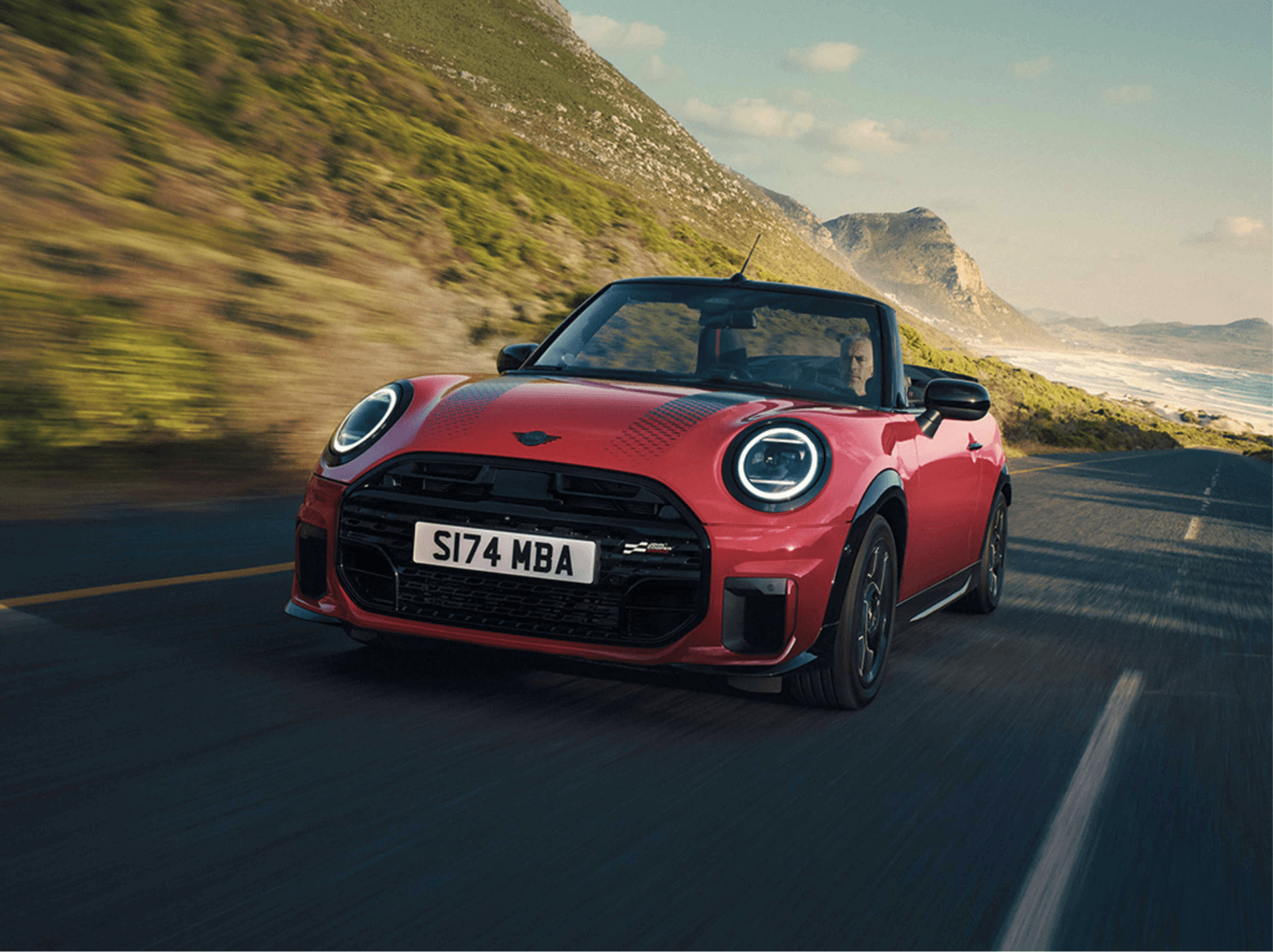 Ein roter Mini Cooper Cabriolet auf einer Küstenstraße. Im Hintergrund befinden sich Berge und das Meer. Das Auto scheint in Bewegung zu sein, was durch die Bewegungsunschärfe im Hintergrund verstärkt wird. Der Fahrer ist im Auto zu sehen.