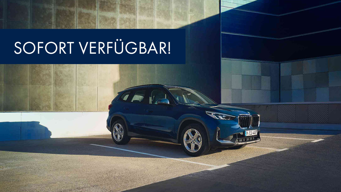 BMW X1 (U11) - Auf einem Parkplatz, die Plätze links und rechts sind frei, in einer statischen 3/4-Frontansicht. Im Hintergrund ist ein Gebäude mit Türen zu sehen. Die Markierungen sind zu erkennen.